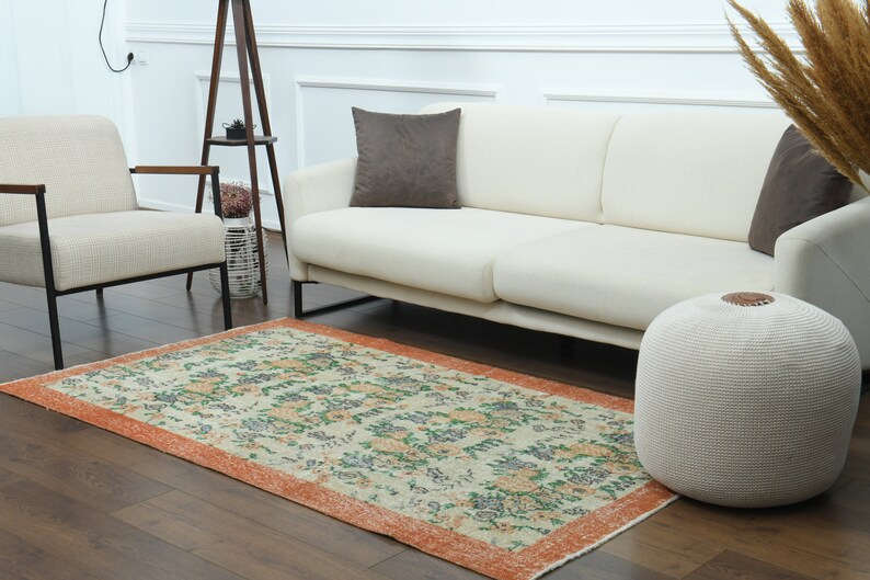 K&ouml;nnte beinhalten: Ein wei&szlig;es Sofa mit einem braunen Kissen steht auf einem Holzboden mit einem gemusterten Teppich. Ein wei&szlig;er Strickhocker steht neben dem Sofa.