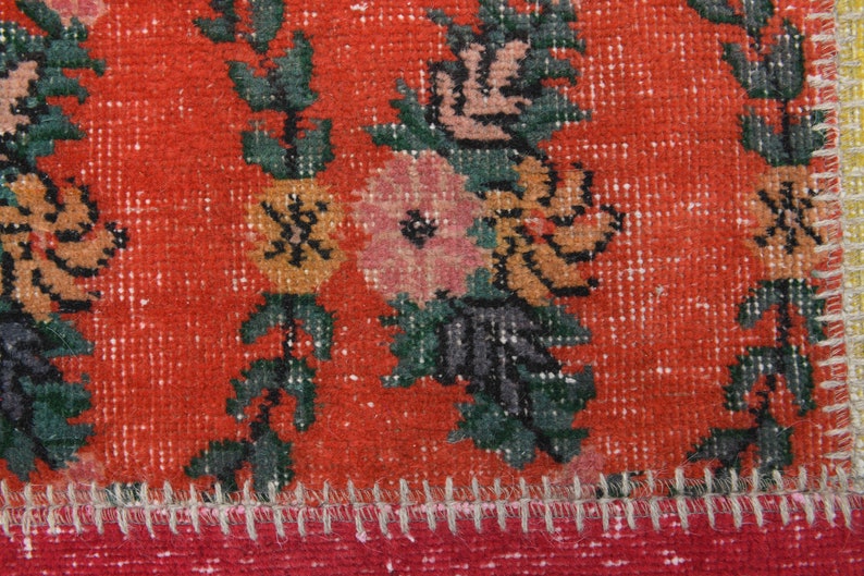 K&ouml;nnte beinhalten: Nahaufnahme eines Abschnitts eines lebendigen, abgenutzten Teppichs. Der Teppich hat einen kr&auml;ftigen roten Hintergrund mit Blumenmustern in Rosa-, Orange- und Gr&uuml;nt&ouml;nen. Die R&auml;nder sind mit einem roten und gelben Besatz versehen, und die N&auml;hte sind sichtbar.