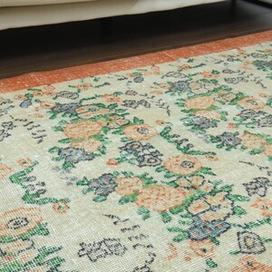 K&ouml;nnte beinhalten: Ein Vintage-Teppich mit floralem Muster auf cremefarbenem Hintergrund und orangefarbenen, gr&uuml;nen und blauen Akzenten. Der Teppich hat einen abgenutzten Look und ist perfekt, um jedem Raum einen Hauch von Vintage-Charme zu verleihen.