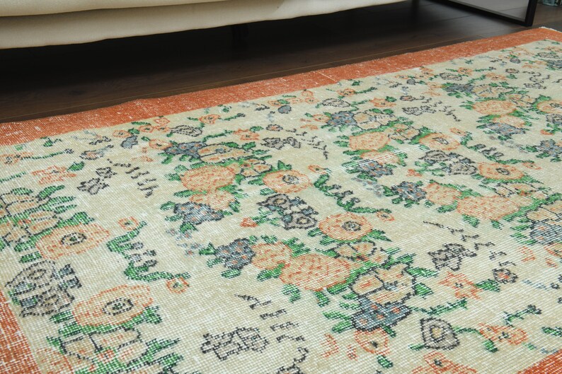 K&ouml;nnte beinhalten: Ein Vintage-Teppich mit floralem Muster auf cremefarbenem Hintergrund und orangefarbenen, gr&uuml;nen und blauen Akzenten. Der Teppich hat einen abgenutzten Look und ist perfekt, um jedem Raum einen Hauch von Vintage-Charme zu verleihen.