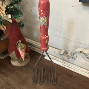 May include: A vintage potato masher with a red, distressed wooden handle and a metal mashing head. The handle shows signs of wear, with paint chipping off to reveal the wood underneath. The masher is a kitchen utensil, likely used for preparing mashed potatoes or other soft foods.
