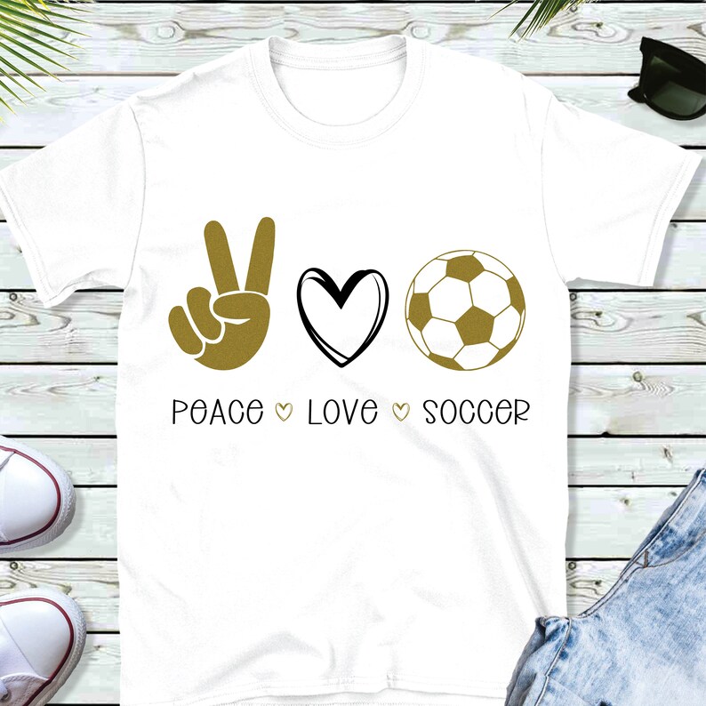 May include: White t-shirt with a gold peace sign, heart, and soccer ball graphic. The text "Peace Love Soccer" is printed below the graphic.