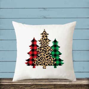 May include: White throw pillow with three Christmas trees printed on it. The trees are in a red and black plaid pattern, a leopard print pattern, and a green and black plaid pattern.
