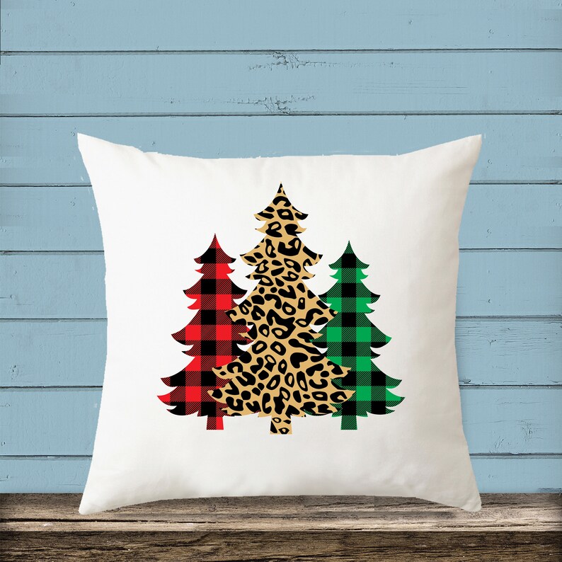 May include: White throw pillow with three Christmas trees printed on it. The trees are in a red and black plaid pattern, a leopard print pattern, and a green and black plaid pattern.