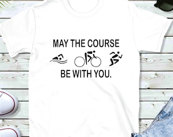 Puede incluir: Camiseta blanca con texto negro que dice "Que el recorrido te acompañe." El texto está acompañado de tres iconos que representan la natación, el ciclismo y la carrera.