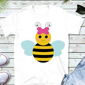 Puede incluir: Camiseta blanca con una abeja de dibujos animados que lleva un lazo rosa. La abeja tiene rayas negras y amarillas, alas azules y una cara sonriente.