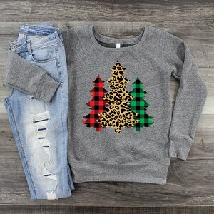 May include: A gray sweatshirt with a design of three Christmas trees. The trees are decorated with red and black buffalo plaid, green and black buffalo plaid, and leopard print.