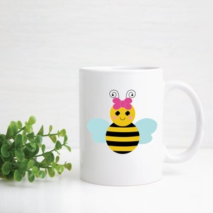 Puede incluir: Taza de cer&aacute;mica blanca con un dise&ntilde;o de abeja de dibujos animados. La abeja es amarilla y negra con un lazo rosa y alas azules.