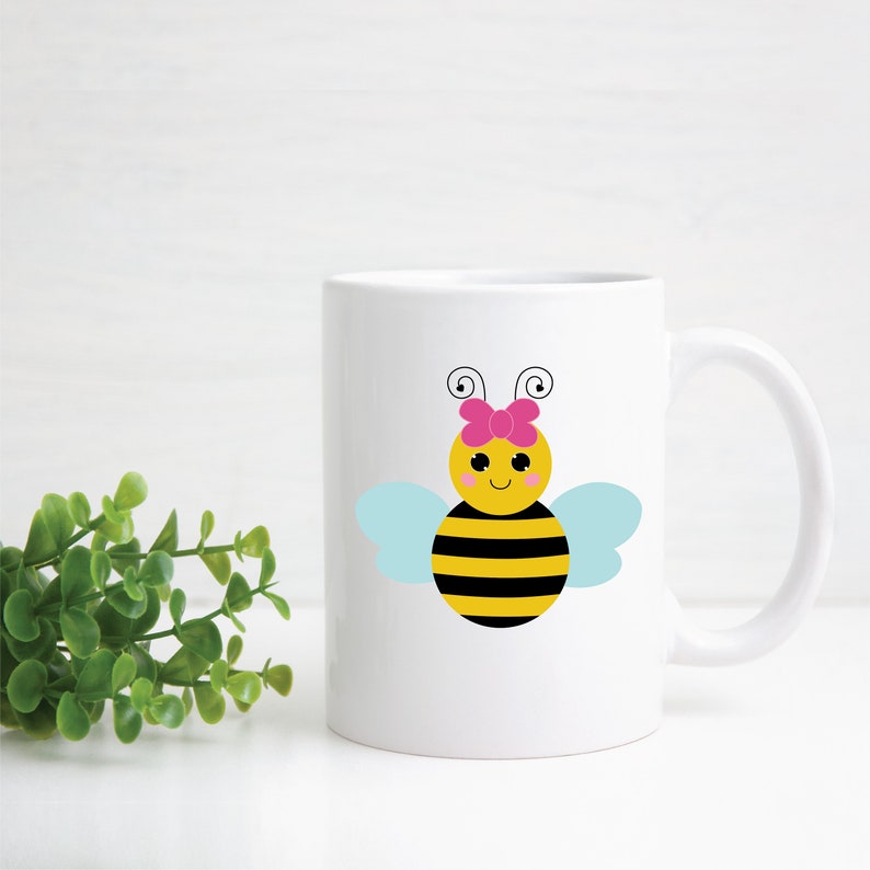Puede incluir: Taza de cer&aacute;mica blanca con un dise&ntilde;o de abeja de dibujos animados. La abeja es amarilla y negra con un lazo rosa y alas azules.