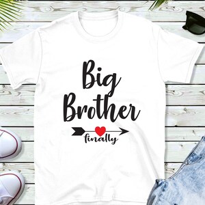 Peut inclure: T-shirt blanc avec un texte noir qui dit "Grand frère enfin" avec un cœur rouge et une flèche.