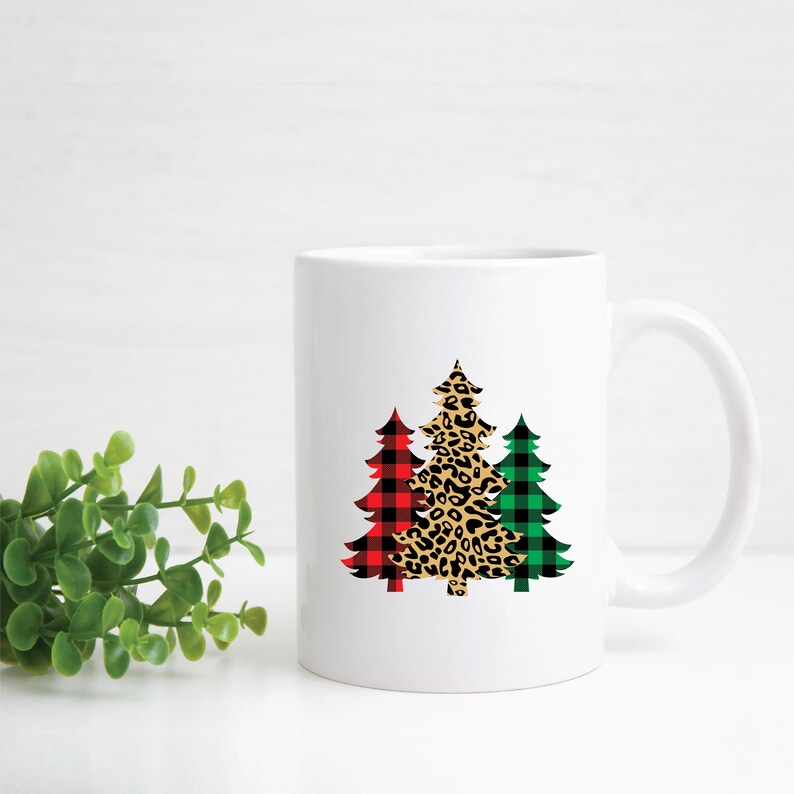 May include: White ceramic mug with a design of three Christmas trees. The trees are decorated with red and black buffalo plaid, leopard print, and green and black buffalo plaid.