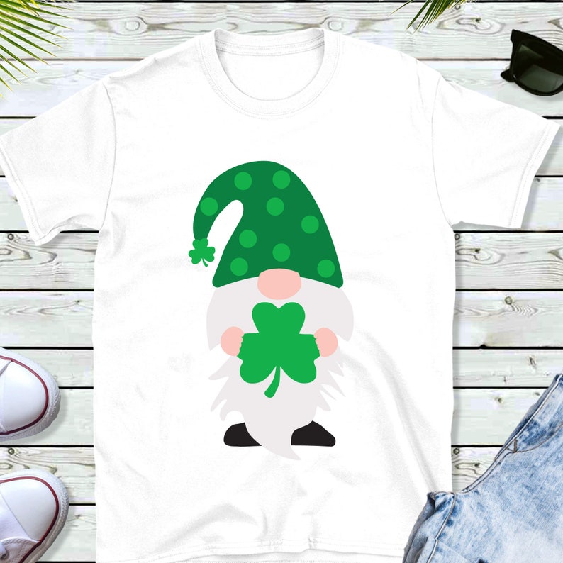 May include: White t-shirt with a green gnome holding a green shamrock. The gnome has a green hat with white polka dots.