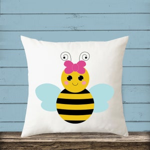 Puede incluir: Almohada blanca con una abeja de dibujos animados que lleva un lazo rosa. La abeja tiene rayas negras y amarillas, alas azules y una cara sonriente.
