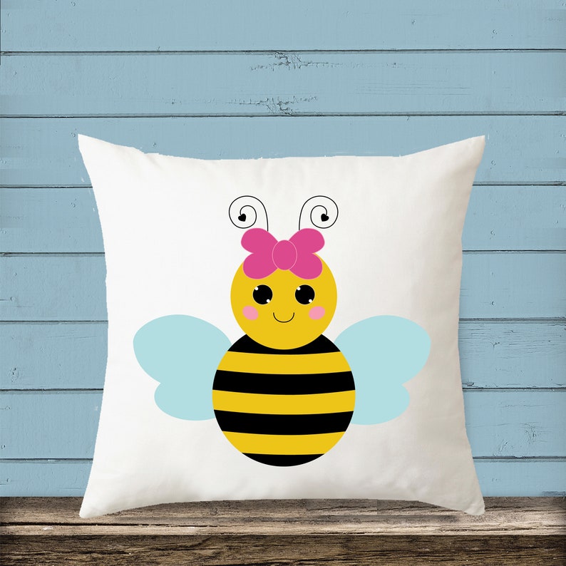 Puede incluir: Almohada blanca con una abeja de dibujos animados que lleva un lazo rosa. La abeja tiene rayas negras y amarillas, alas azules y una cara sonriente.