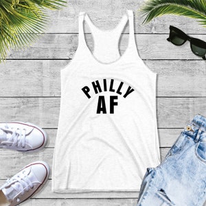 Puede incluir: Camiseta blanca de tirantes con un gr&aacute;fico negro que dice "Philly AF" sobre un fondo de madera blanca.