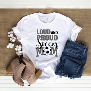 May include: White t-shirt with the text "Loud and Proud Soccer Mom" in black and gray lettering with a soccer ball graphic.