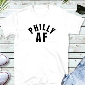 Puede incluir: Camiseta blanca con texto negro que dice "Philly AF" en arco.