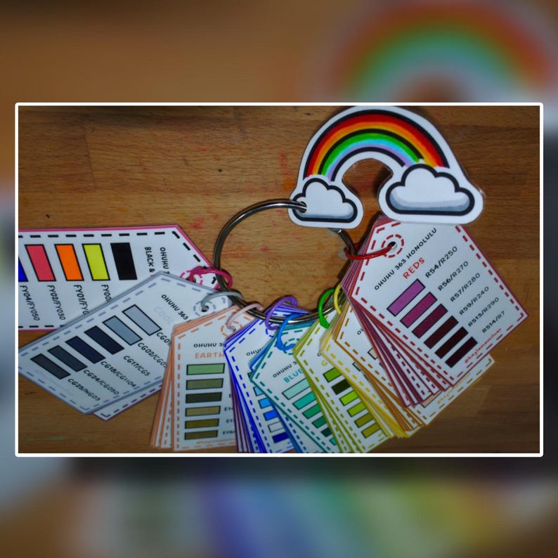 Puede incluir: Un llavero de muestras de color con varias paletas, incluyendo rojos, azules y tonos tierra. Cada muestra est&aacute; etiquetada con c&oacute;digos y nombres de color. Un adorno en forma de arco&iacute;ris con nubes corona el llavero, a&ntilde;adiendo un toque caprichoso.