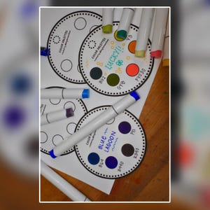 K&ouml;nnte beinhalten: Draufsicht auf wei&szlig;e Kunstmarker und Farbkarten. Die Karten sind kreisf&ouml;rmig mit Farbfeldern und Text wie "Blue Lagoon" und "Lucky!!". Marker haben farbige Spitzen, darunter Blau, Lila und Gelb.