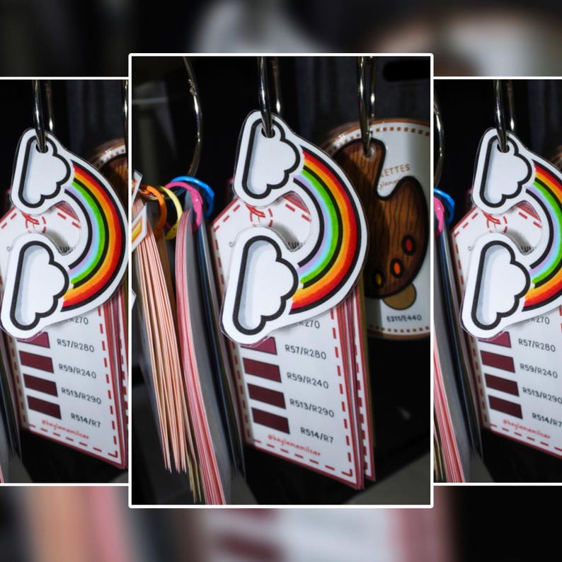 Puede incluir: Un conjunto de etiquetas coloridas con un dise&ntilde;o de arco&iacute;ris y nubes. Cada etiqueta, unida a un anillo met&aacute;lico, presenta muestras de color y texto. Las etiquetas est&aacute;n colgadas en una exhibici&oacute;n.