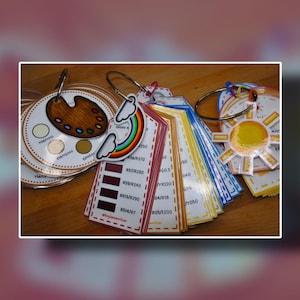 Puede incluir: Una colección de tarjetas de muestras de color en anillos de metal. Las tarjetas muestran varios tonos de color, incluyendo rojos, amarillos, azules y marrones. Algunas tarjetas tienen etiquetas de texto con códigos y nombres de color. Una tarjeta presenta un encanto de arcoíris y nube.