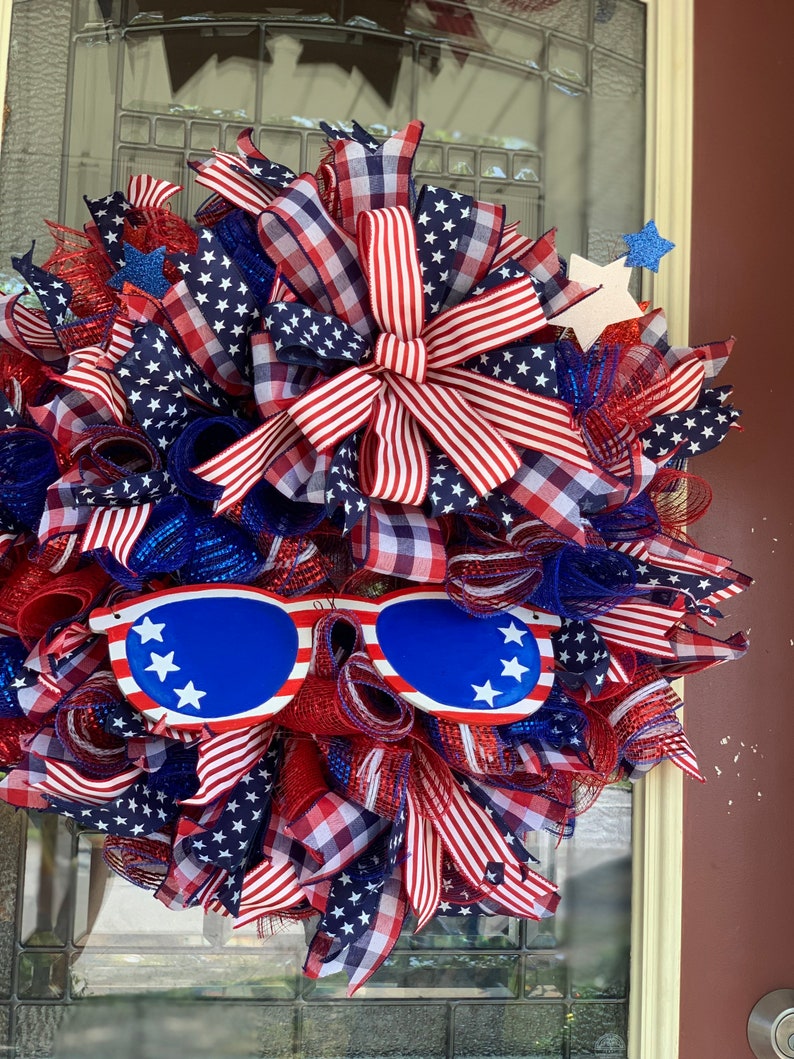 May include: A red, white, and blue patriotic wreath with a large bow, stars, and a pair of blue sunglasses with white stars.