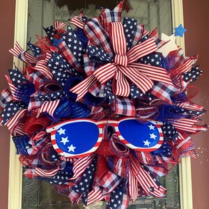 May include: A red, white, and blue patriotic wreath with a large bow and sunglasses. The wreath is decorated with stars and stripes.