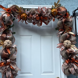 May include: A decorative door garland featuring metallic copper and brown mesh ribbons, fabric pumpkins, and autumn-themed foliage. A small wooden sign with the text hello fall is integrated into the arrangement, which frames a white door.
