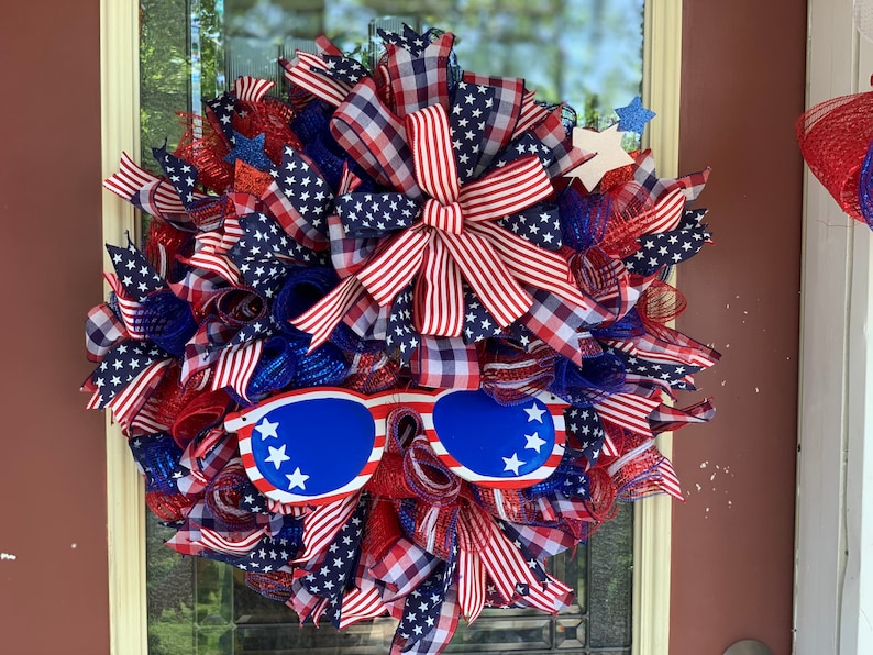 May include: A red, white, and blue patriotic wreath with a star-spangled ribbon bow and a pair of blue sunglasses with white stars. The wreath is decorated with red, white, and blue mesh and ribbon.