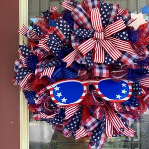 May include: A red, white, and blue patriotic wreath with a red, white, and blue checkered bow and a pair of blue sunglasses with white stars.