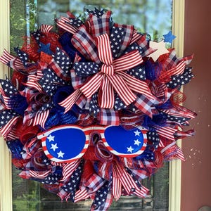 May include: A red, white, and blue patriotic wreath with a large bow and sunglasses. The wreath is decorated with stars and stripes.
