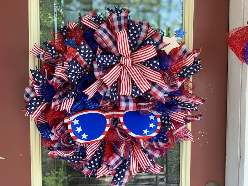 May include: A red, white, and blue patriotic wreath with a large bow and sunglasses. The wreath is decorated with stars and stripes.