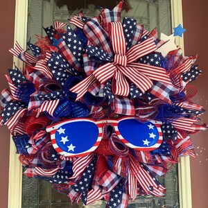 May include: A red, white, and blue patriotic wreath with a large bow and sunglasses. The wreath is decorated with stars and stripes.