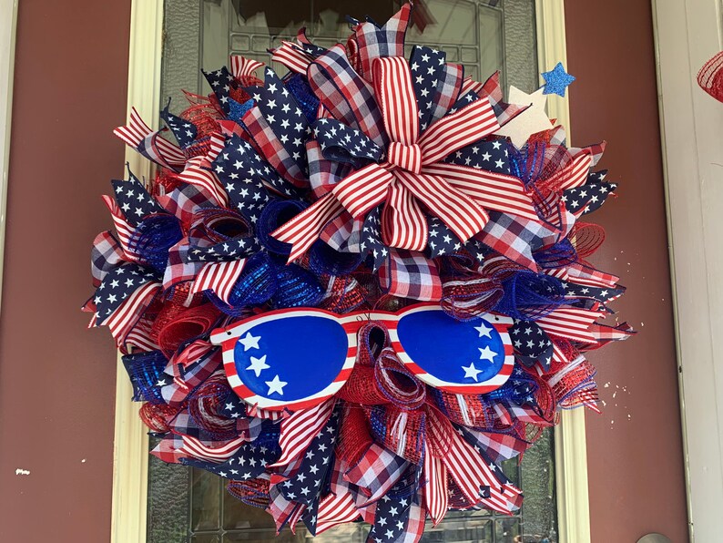 May include: A red, white, and blue patriotic wreath with a large bow and sunglasses. The wreath is decorated with stars and stripes.
