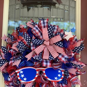 May include: A red, white, and blue patriotic wreath with a large bow and sunglasses. The wreath is made of mesh and ribbon and features stars and stripes.