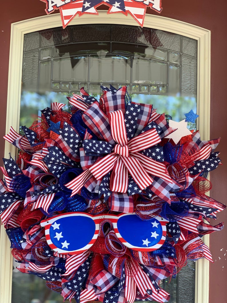 May include: A red, white, and blue patriotic wreath with a large bow and sunglasses. The wreath is made of mesh and ribbon and features stars and stripes.