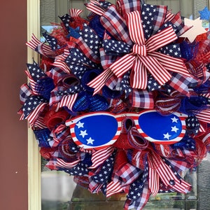 May include: A red, white, and blue patriotic wreath with a large bow and sunglasses. The wreath is decorated with stars and stripes.