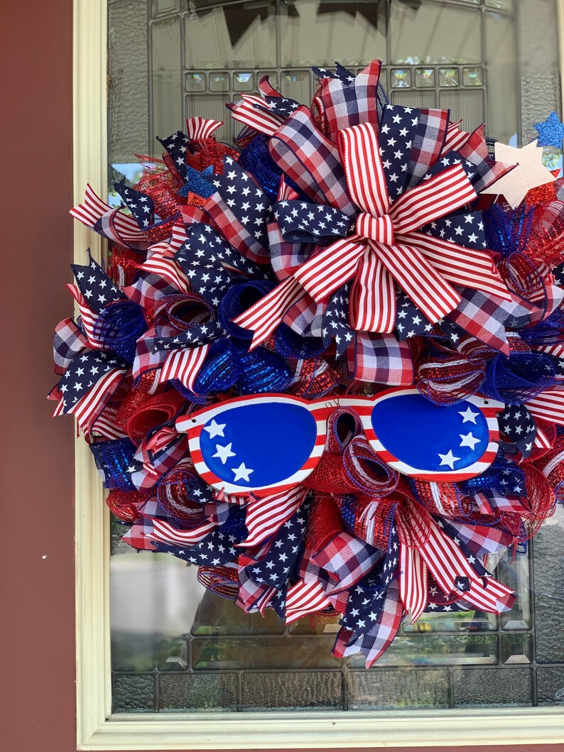 May include: A red, white, and blue patriotic wreath with a large bow and sunglasses. The wreath is decorated with stars and stripes.