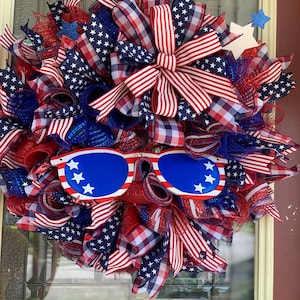 May include: A red, white, and blue patriotic wreath with a red, white, and blue bow and a pair of blue sunglasses with white stars. The wreath is made of mesh and ribbon and is decorated with stars and stripes.