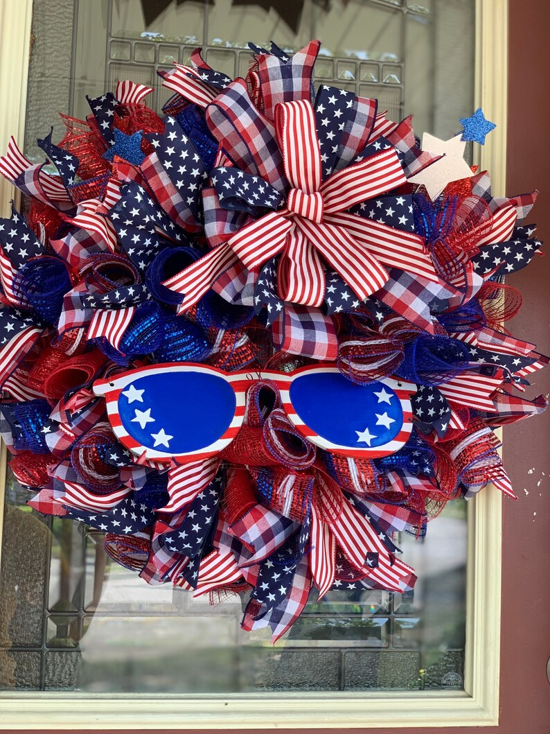 May include: A red, white, and blue patriotic wreath with a red, white, and blue bow and a pair of blue sunglasses with white stars. The wreath is made of mesh and ribbon and is decorated with stars and stripes.