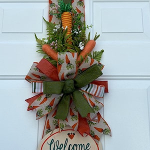 May include: A carrot-themed door decoration with a large bow made of orange and green ribbons, artificial carrots, and foliage. The sign says "Welcome to our Carrot Patch Open Daily."