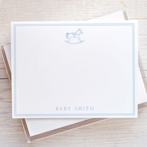 May include: A stack of white note cards with a blue border and a rocking horse illustration. The cards have the text "BABY SMITH" printed on the bottom.