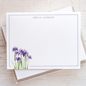 May include: White stationery with a purple border and watercolor irises in the corner. The name "AMELIA JOHNSON" is printed in purple at the top. The stationery is displayed in a clear plastic box.