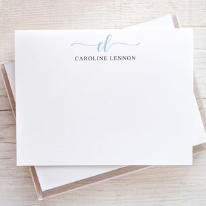 May include: White stationery with a light blue cursive "cl" and the name "Caroline Lennon" printed in black. The stationery is stacked in a clear plastic case, set against a light wood grain background.