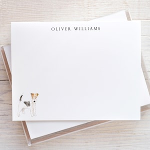 May include: White stationery set featuring the name "OLIVER WILLIAMS" in black font. A watercolor illustration of a dog is in the lower left corner. The stationery is stacked in a clear plastic box, resting on a light wood surface.