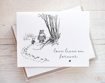 May include: A white greeting card with a black and white illustration of a bear and pig walking on a path. The text "love lives on forever" is written in cursive with a small heart below.