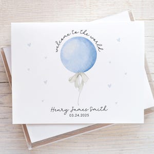 May include: A white card with a watercolor blue balloon design and the text "welcome to the world." The card includes the name "Henry James Smith" and the date "03.24.2025." Small gray hearts are scattered around the balloon.