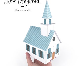 May include: A paper model of a white New England church with a blue roof and steeple. The model is held in a hand, and the text "New England Church model" is visible above the model.