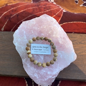 May include: A beaded bracelet made of Picture Jasper gemstones, resting on a large pink crystal. The bracelet features round, earth-toned beads. A small card reads "Gems in this Design - Picture Jasper - harmony - creative visualization."