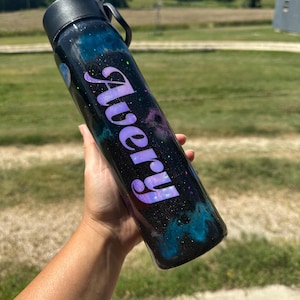 Puede incluir: Botella de agua negra con diseño de galaxia, con detalles en azul, morado y blanco. El nombre "Avery" está escrito en púrpura. La botella tiene tapa negra y asa.